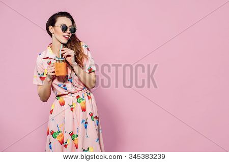 Beautiful Woman In Dress With Printed Friuts Posing With Glass Of Fresh Friut Drink On Pink Backgrou