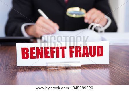 Close-up Of A Nameplate With Benefit Fraud Title With Man Checking Documents Using Magnifying Glass