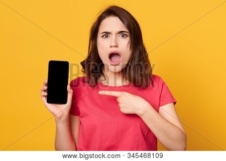 Portrait Of Good Looking Young Brunette Female Showing Smartphone Screen And Points At It With Her F