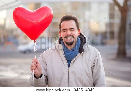Portrait Of Smiling Man Standing With Closed Eyes And Holding With  Red Air Balloon Shaped Like Hear