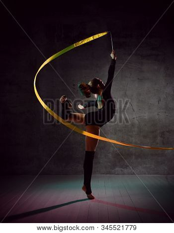 Young Girl Gymnast In Black Sport Body And Uppers Standing With Streched Leg And Making Exercise Wit