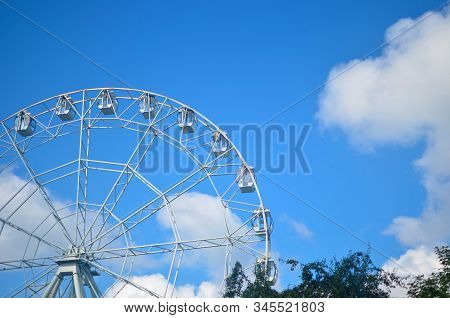 White Ferris Wheel Carousel On A Background Of Blue Blue Sky With White Clouds And Green Foliage On 