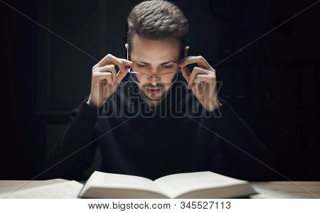 Young Bearded Man Holding Glasses Reading Book On Dark Background