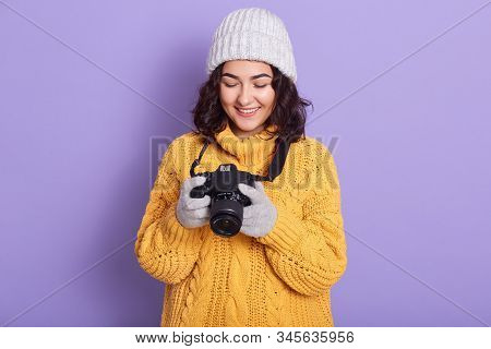 Hirizontal Shot Of Beautiful Young Girl Wearing Cap, Knitted Sweater Holding Camera And Looking At D