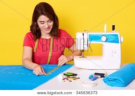 Horozontal Shot Of Smiling Young Woman Sewing With Professional Machine At Workshop, Taking Measurme