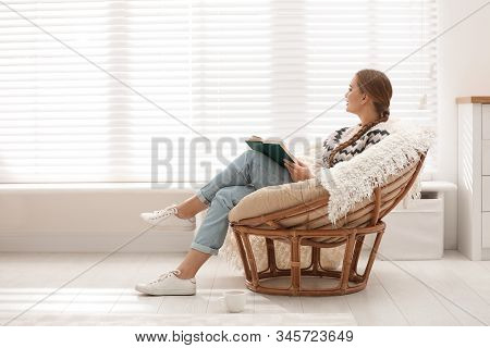 Young Woman Reading Book In Papasan Chair Near Window At Home