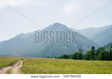 Сountry Winding Dirt Road Leads Into Giant Mountain With Conifer Forest In Haze. Alpine Scenic Lands