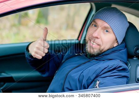 Warmly Dressed Young Smiling Bearded Charismatic Man Sitting Behind The Wheel Of A Car With A Raised