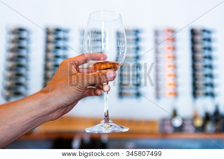 Man Hand Holding A Glass Of White Wine In Selective Focus View, Wine Bottles On Wall Storage Racks A