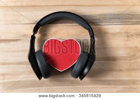 Full-size Wireless Headphones Pulled Over Small Red Heart-shaped Box On A Light Brown Wooden Table. 