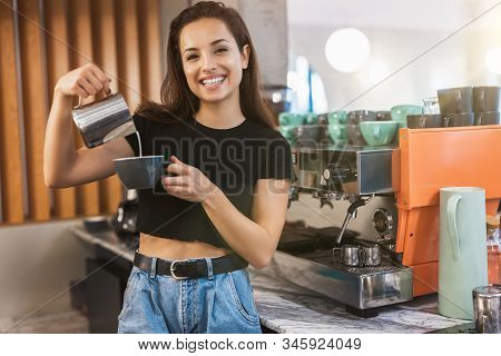 Young Smiling Barista Woman Pouring Whipped Milk Into Coffee Drink, Preparing Latte Macchiato, Looki