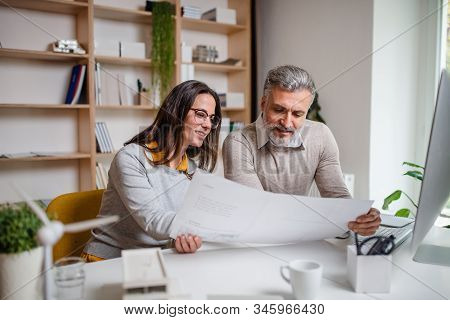Architects Sitting At The Desk Indoors In Office, Looking At Blueprints.