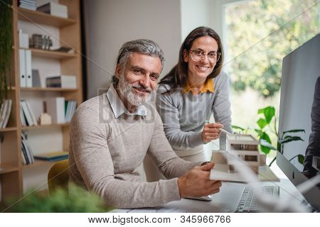 Architects With Model Of House Sitting At The Desk Indoors In Office, Working.