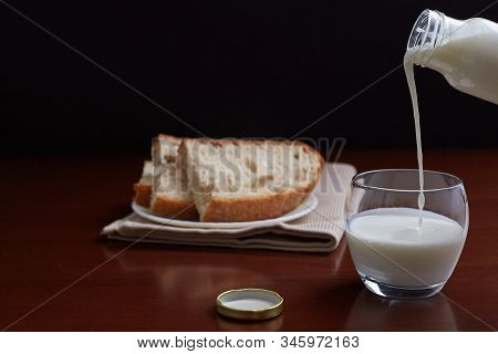 Kefir From A Bottle Flows Into A Glass. Bread In The Background. Dark Background And Brown Wooden Ta