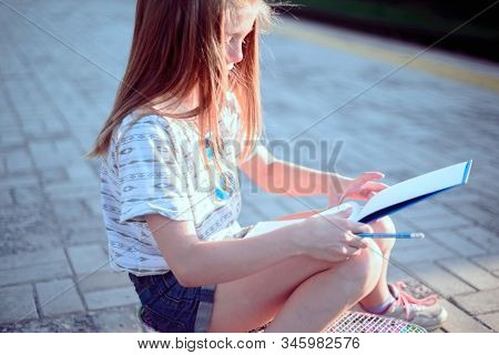Beautiful Little Girl Sitting On A Skateboard And Writing In A Tetrad. Outdoor Lifestyle