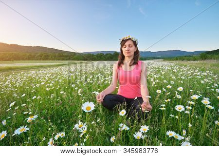 Girl meditates in lotus pose on green camomile meadow. Practicing of yoga at summer. Portrait of young peaceful woman. Training and meditation outdoor. Healthy lifestyle.