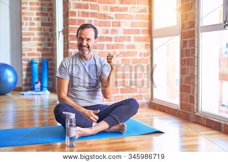 Middle age handsome sportman sitting on mat doing stretching yoga exercise at gym smiling with happy face looking and pointing to the side with thumb up.