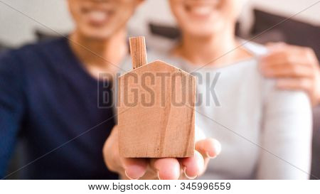 Home Loan And Financial Concept - Close Up Wooden Home Toy In Hand With Blurred Asian Couple Smiling