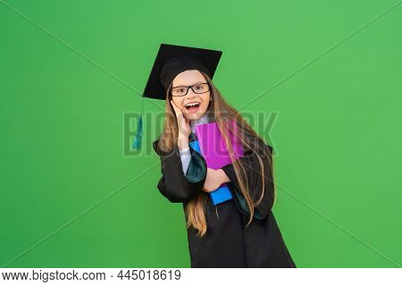 A Beautiful Little Schoolgirl In A Graduation Cap And A Ceremonial Robe, Holding Books On An Isolate
