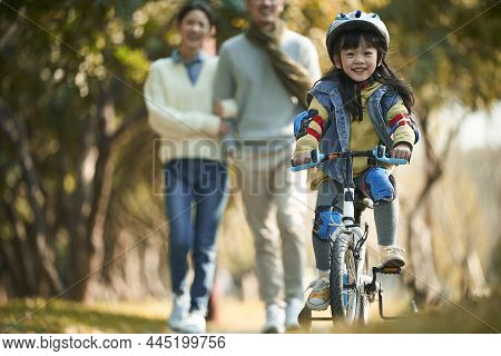 Little Asian Girl With Helmet And Full Protection Gears Riding Bike In City Park With Parents Watchi