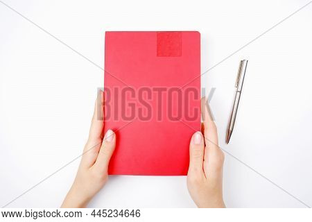 The Girl Holds A Red Diary Or Notebook Next To A Gray Metal Pen On A White Background.the Concept Of