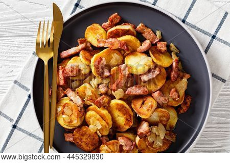 Bratkartoffeln, Fried Potatoes With Bacon And Onion On A Plate With Cutlery On A White Wooden Table,