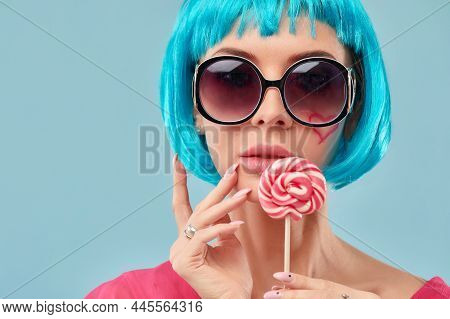 Young Funky Girl In Blue Wig And Eyeglasses, Holding Lollipop Candy, Looking Aside With Dreamful Smi