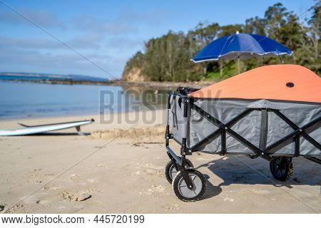 Outdoor Beach Cart Wagon, Beach Umbrella An Stand Up Paddle Board On A Sandy Beach Near The Ocean. F