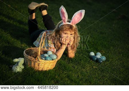 Cute Bunny Child With Rabbit Ears. Child With Easter Eggs In Basket Outdoor. Kid Laying On Grass In 