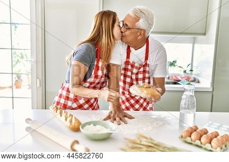 Middle age hispanic couple kising and cooking bread at the kitchen.