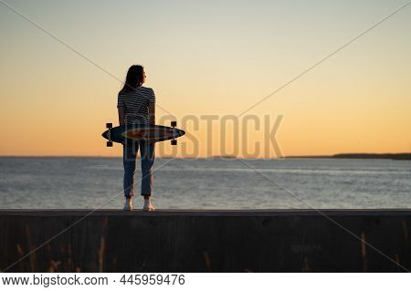Young Fashion Girl With Longboard Enjoy Sunset Looking At Seaside Skyline. Stylish Trendy Female Lon