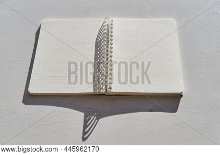 An Open Blank, Checkered And Spiral-bound Notebook Lying On A White Table In Bright Sunlight, With H