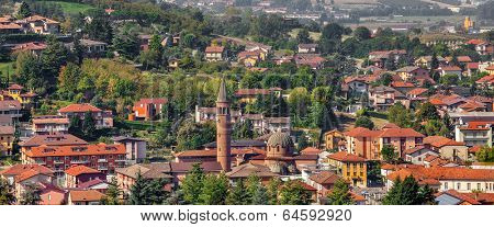 View of church's bell tower among houses and green trees in town of Alba in Piedmont, Northern Italy (panorama).