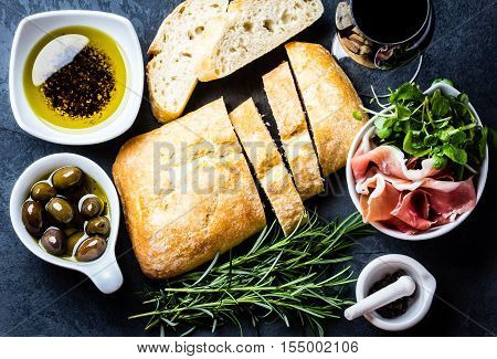Bread ciabatta, jamon ham serrano paleta iberica, arugula, olive pepper oil, olives, rurosemary and glass of red wine on stone slate black background. Top view copy space