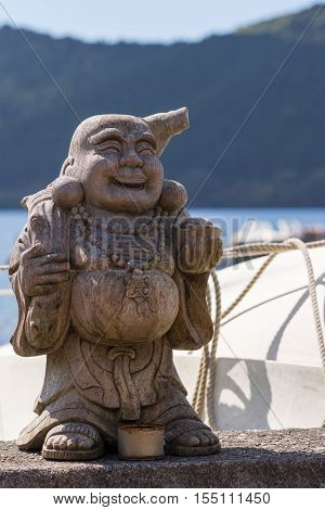 Hakone Japan - September 27 2016: A wooden statue of the laughing and obese Japanese God Ebisu stands at the shoreline of Lake Ashi. White color and rope of boat forest and water.