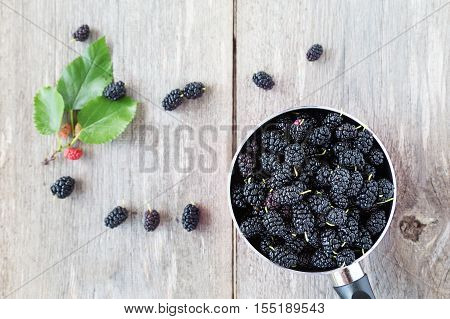 black mulberry berries in a pink pot on a wooden table. in the background are mulberry berries and sprig with ripened berries.