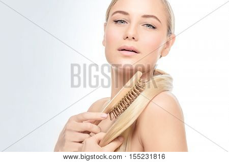 Portrait of a gorgeous woman over clear background combing her blond hair with wooden hairbrush, fashion model wearing natural makeup