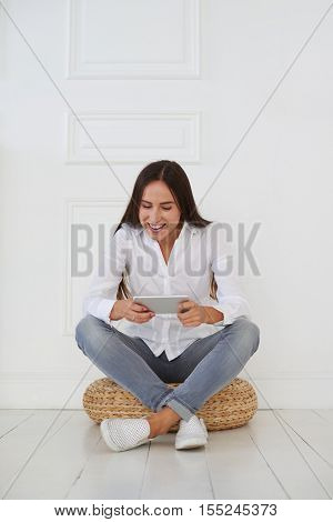 Modern young businesswoman using her tablet with joyful facial expression while sitting on a hassock against white background 