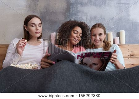Merry weekend. Cute smiling involved friends reading the magazine and eating popcorn while lying on the bed at home and being covered with the blanket