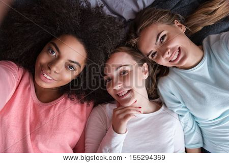 Joyful weekend. Cheerful delighted young girls smiling and lying on the bed while taking selfie and expressing joy in the bedroom