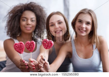 Funny free time . Delighted smiling positive girls holding lollipops and sitting in the bedroom while expressing joy at the home party