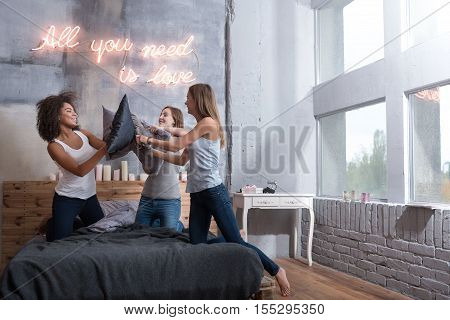 Pillow fight. Delighted amused young girls smiling and standing on the bed in the bedroom while having a funny fight with pillows