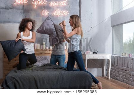 We having fun. Delighted amused young girls smiling and standing on the bed in the bedroom while having a funny pillow fight