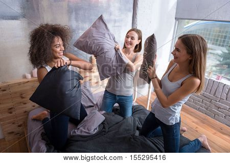 unny fight at home party. Amused laughing young girls standing on the bed in the bedroom while having a funny pillow fight and expressing joy
