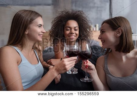 We like wine. Pleasant delighted young girls smiling and sitting in the bedroom while expressing happiness and drinking wine