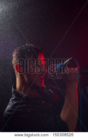 Young attractive male singer performing with microphone on the stage at the background of red spotlights.