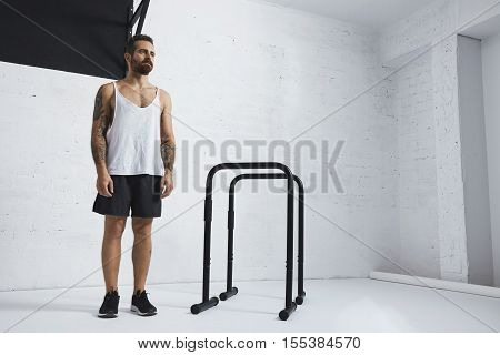 Brutal tattooed bearded male athlete in blank white tank t-shirt standing near to parallel bars and pull bar looking on side , ready to workout
