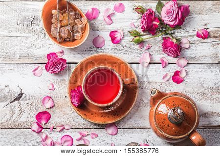 Rose buds tea in a cup, flowers and tea pot on white wooden table, top view.