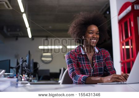 black woman in modern office at her workplace speeking on phone over earphones and working on laptop computer