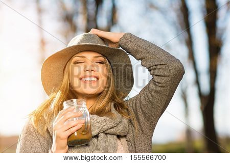 Closeup of gorgeous happy young blonde woman with coffee in park in autumn. Modern beautiful teenage girl in gray outfit and floppy hat smiling, enjoying sunny fall day outside.